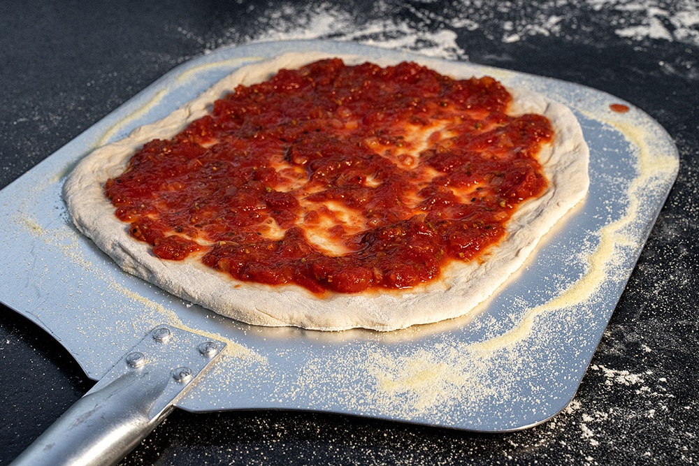 Pizza mit Tomatensauce bestreichen, Pizzaschaufel mit Hartweizengrieß bestreuen, Teig auflegen.