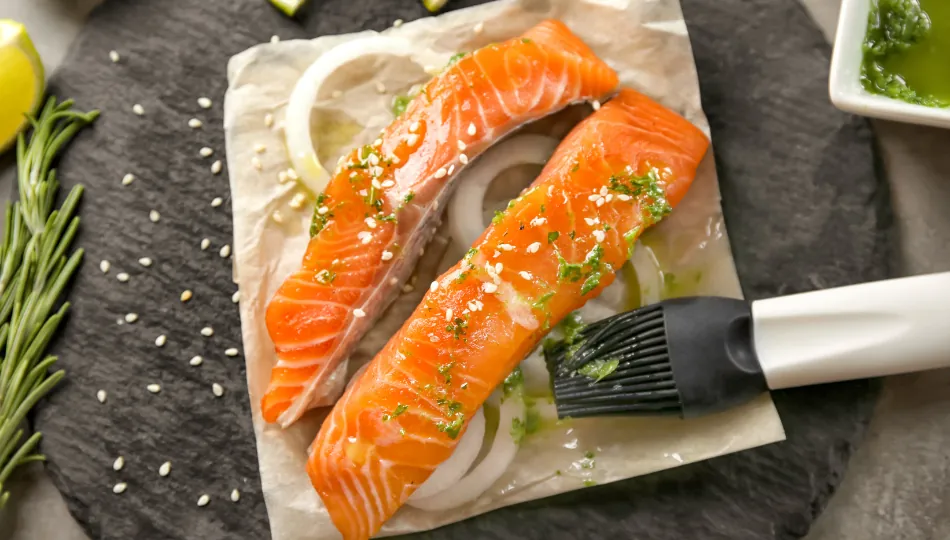 Zwei frische Lachsfilets auf Backpapier, die mit einer grünen Kräutermarinade, Zwiebelringen und Sesam bestreut werden, um den Lachs marinieren zu können.