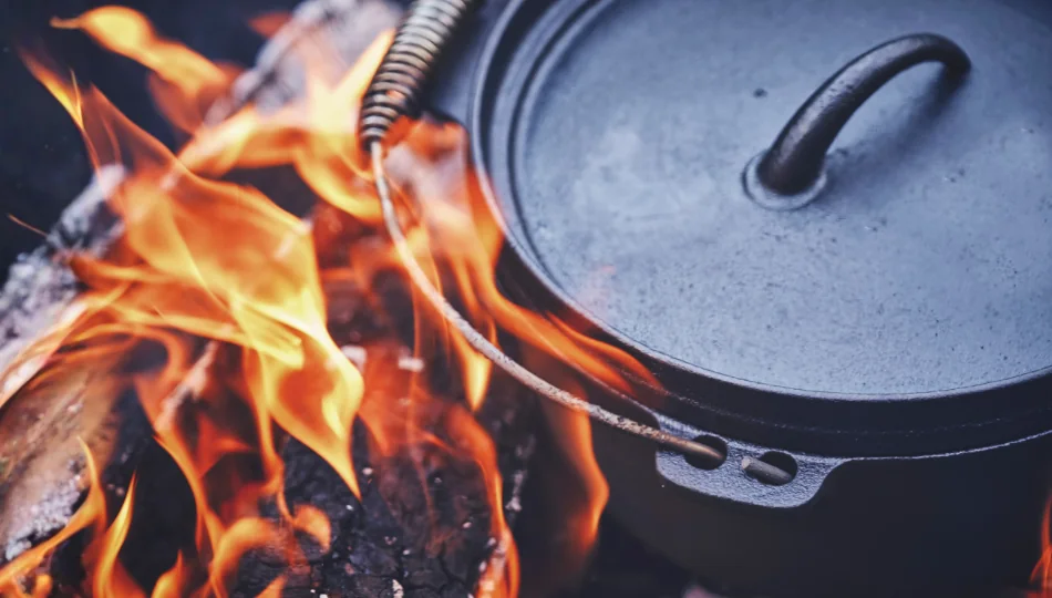 Ein schwarzer, gusseiserner Dutch Oven mit Deckel und einem Griff mit Holzrolle steht über brennenden Holzscheiten in einer runden Metall-Feuerschale. Helle orangefarbene Flammen lodern um den Topf.