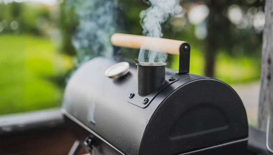 Ein klassischer, schwarzer BBQ-Offset-Smoker im Garten, aus dessen Schornstein leichter Rauch aufsteigt, um zu veranschaulichen: Was ist ein Smoker-Grill und wie funktioniert das langsame Garen mit Holzfeuer